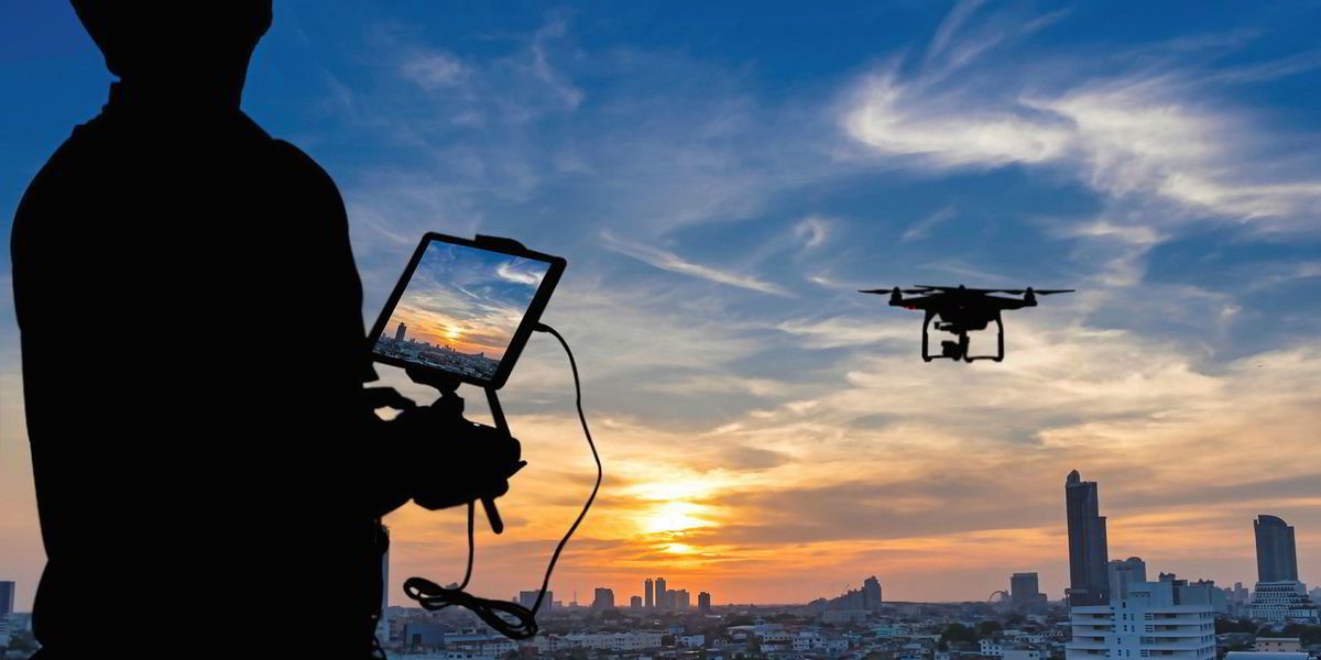 A silhouetted figure overlooking a city skyline at dusk, controlling a drone through a tablet computer