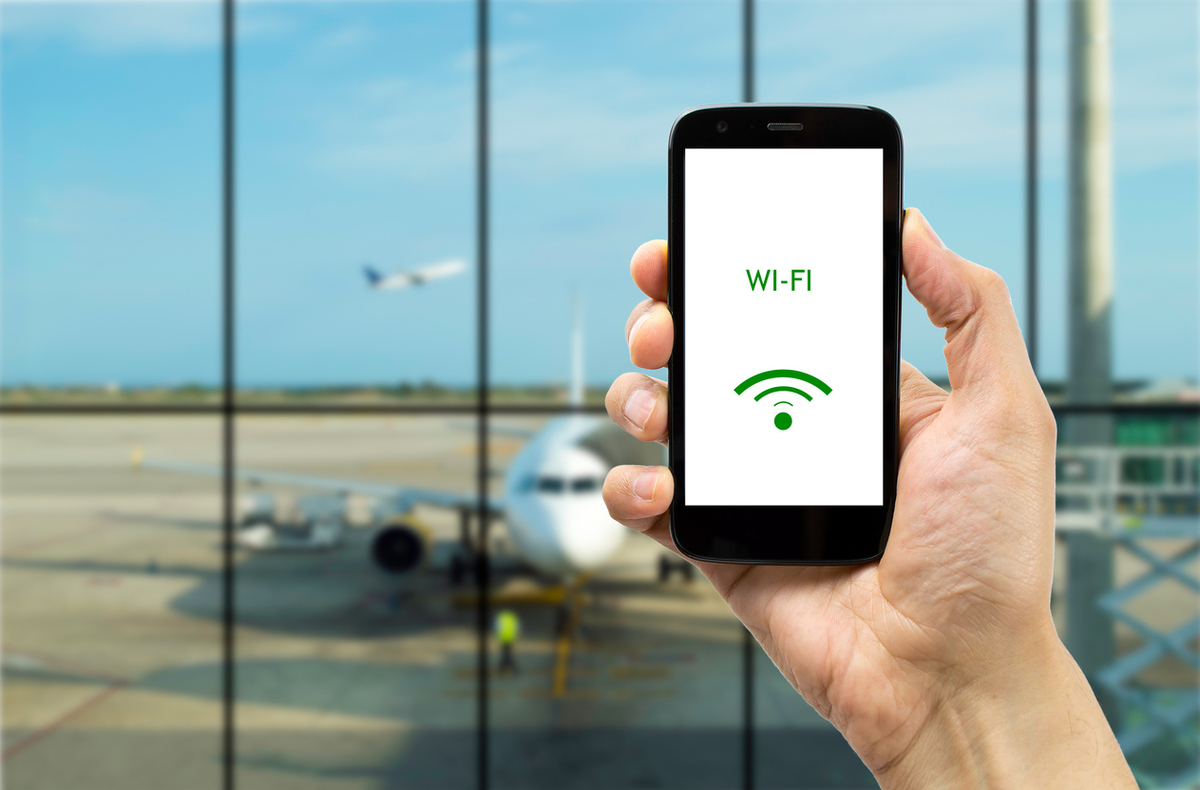 A hand holding a mobile phone displaying a Wi-Fi symbol, in front of a window revealing an airport setting
